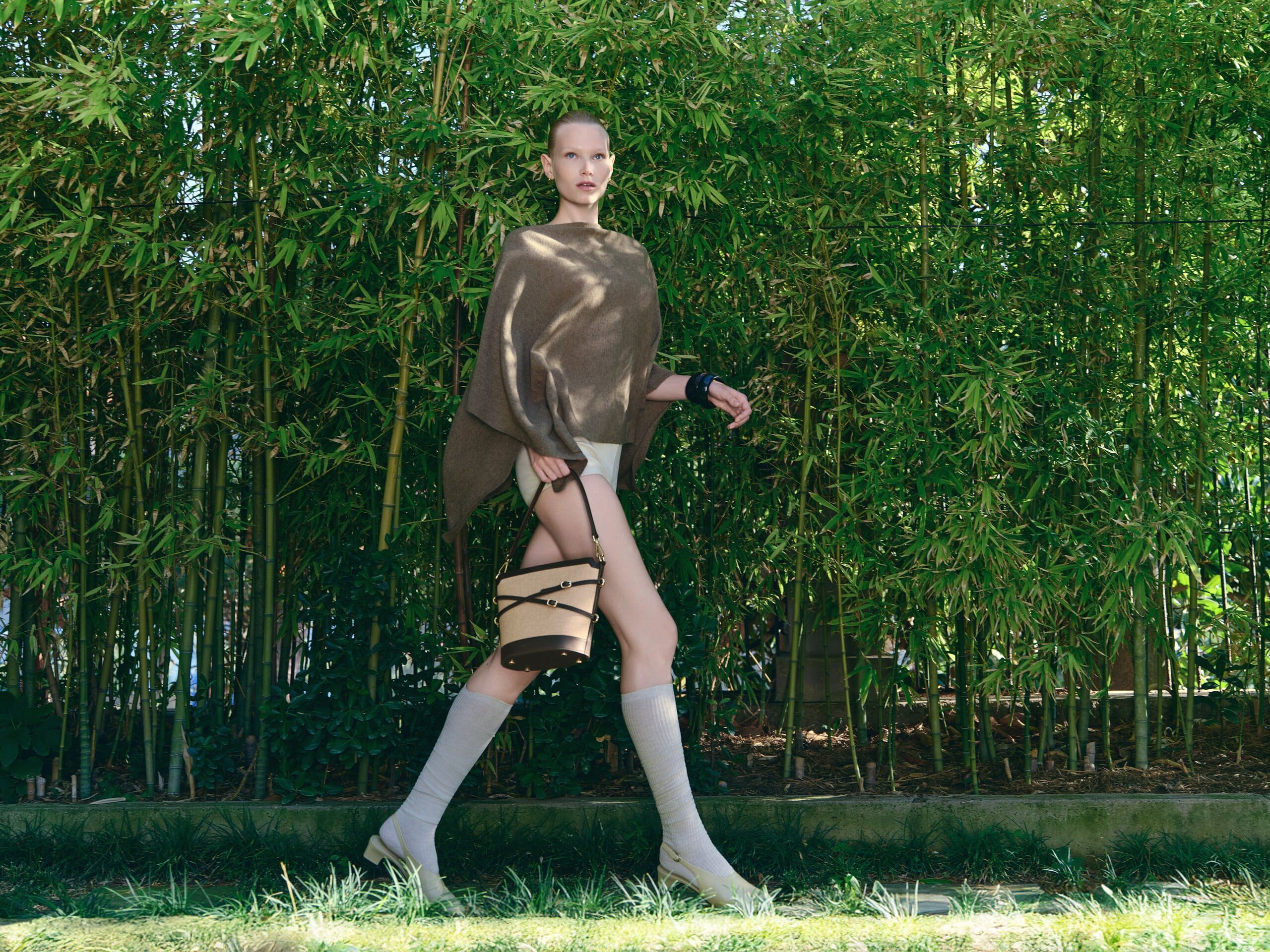 Mid-stride along a bamboo hedge in a taupe poncho, the canvas bucket bag swinging at the knee for Owlish Studio.