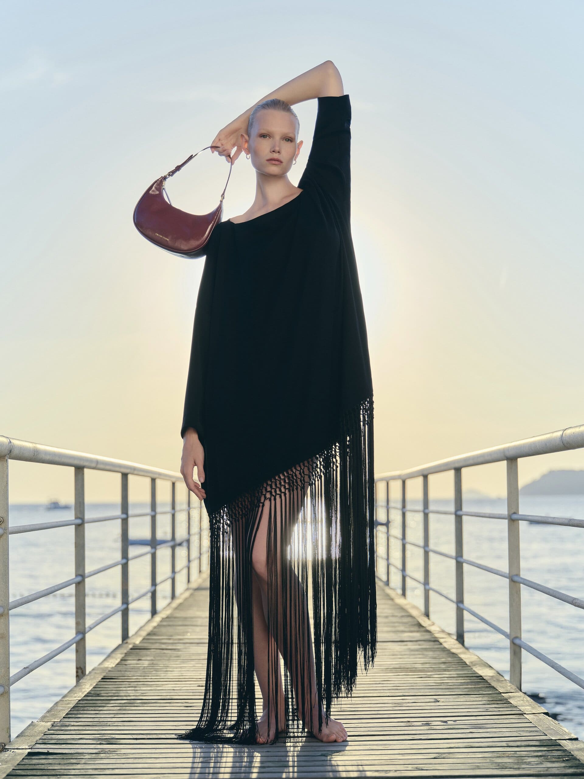 Arm arched against the sunset on a Bosphorus pier, the fringed black dress backlit and the burgundy hobo held aloft for Owlish Studio.