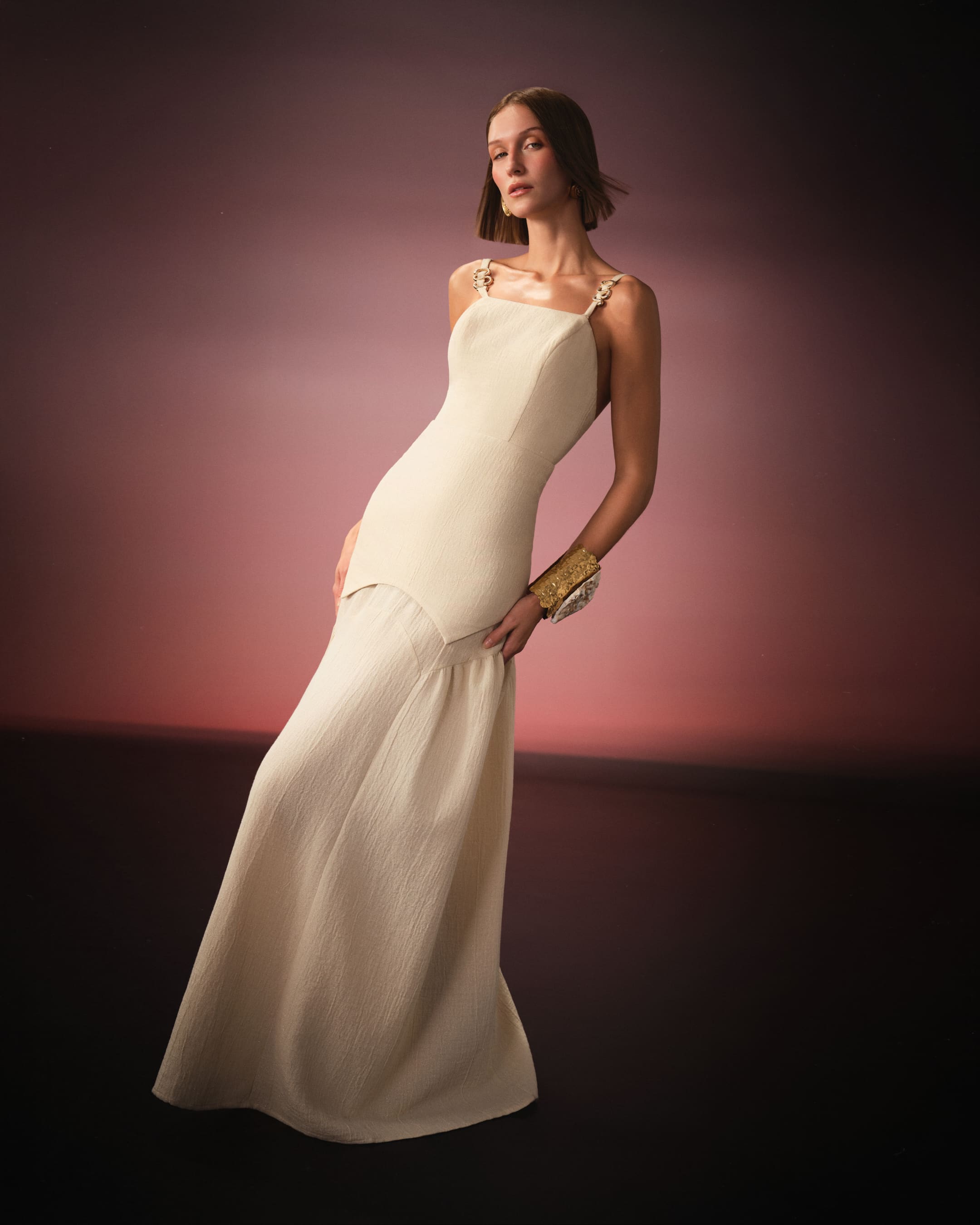 An ivory gown with gold hardware straps leaning into a rose horizon for Saint Ones.