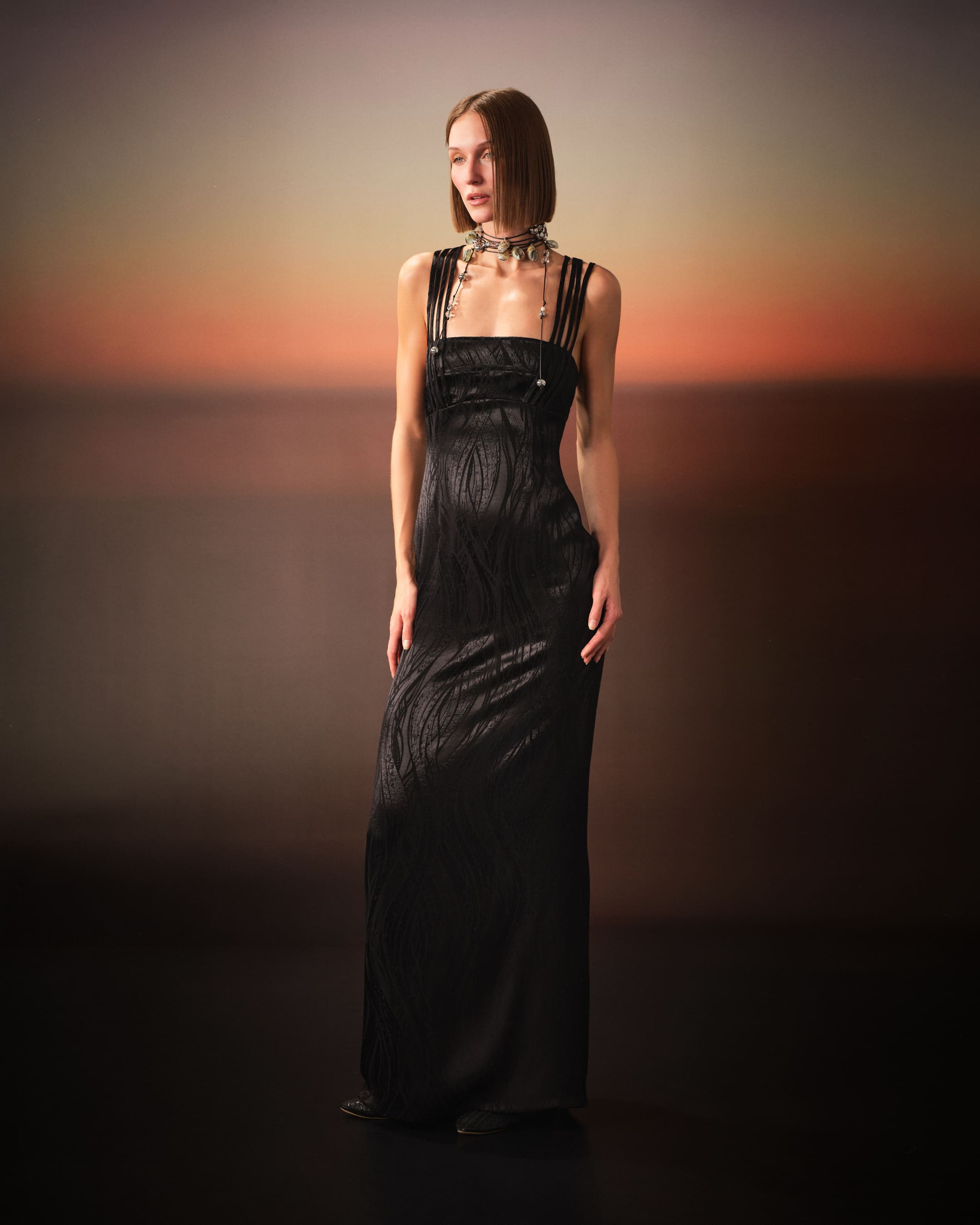 A strap-shouldered black satin gown with a shell lariat under burnt-orange dusk for Saint Ones.