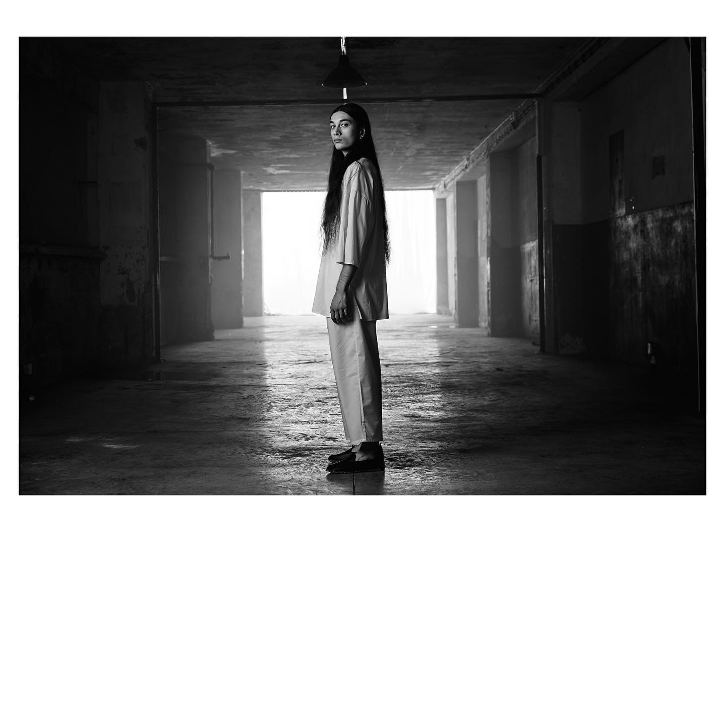 A long-haired figure in loose white poplin stands in profile through a derelict factory hall for ST Studio.