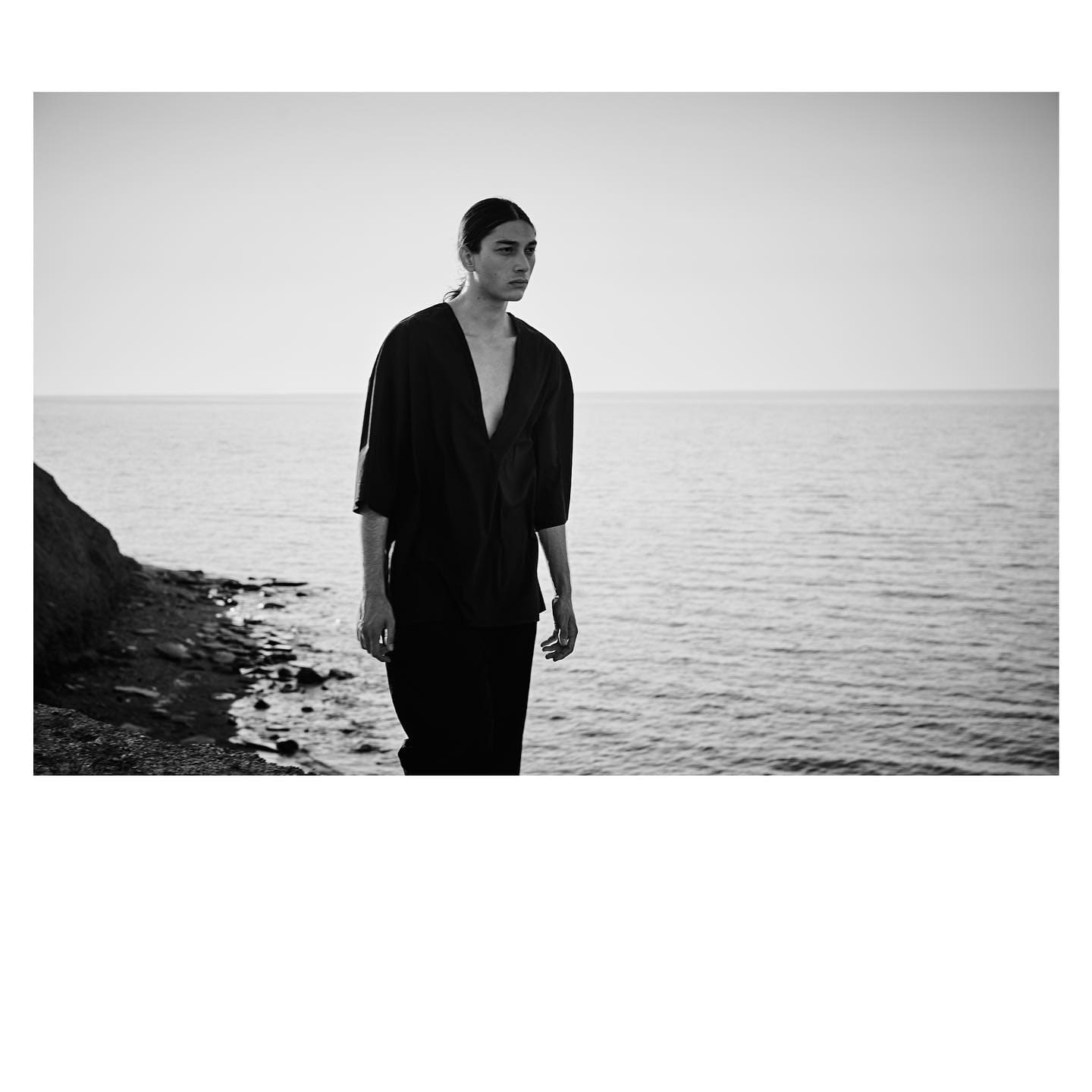 The open black tunic meets a flat evening sea as ST Studio's second face walks the rocky shoreline.