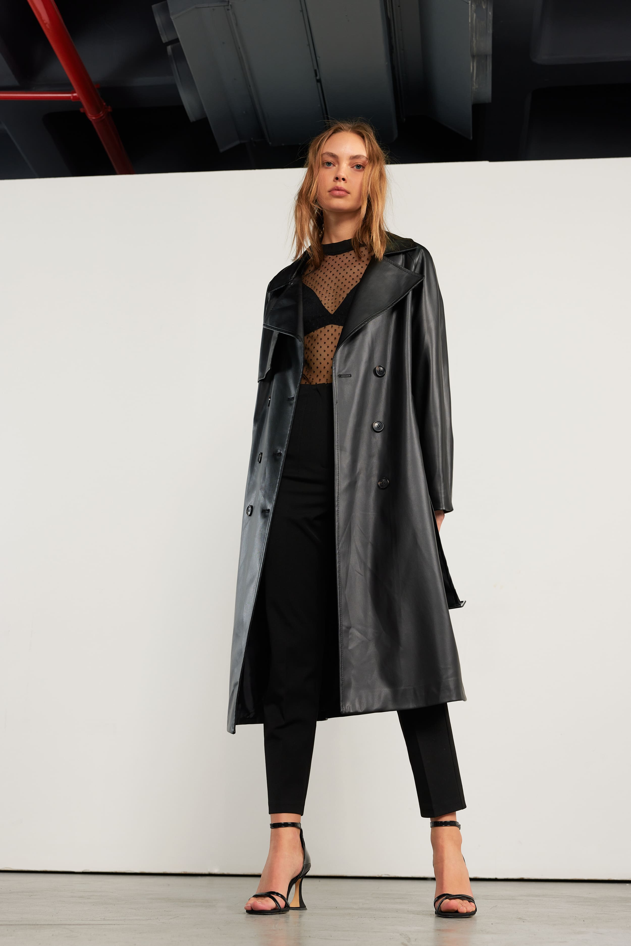 A black faux-leather trench thrown open over dotted tulle, framed by white panel and industrial ceiling for Vatkalı.
