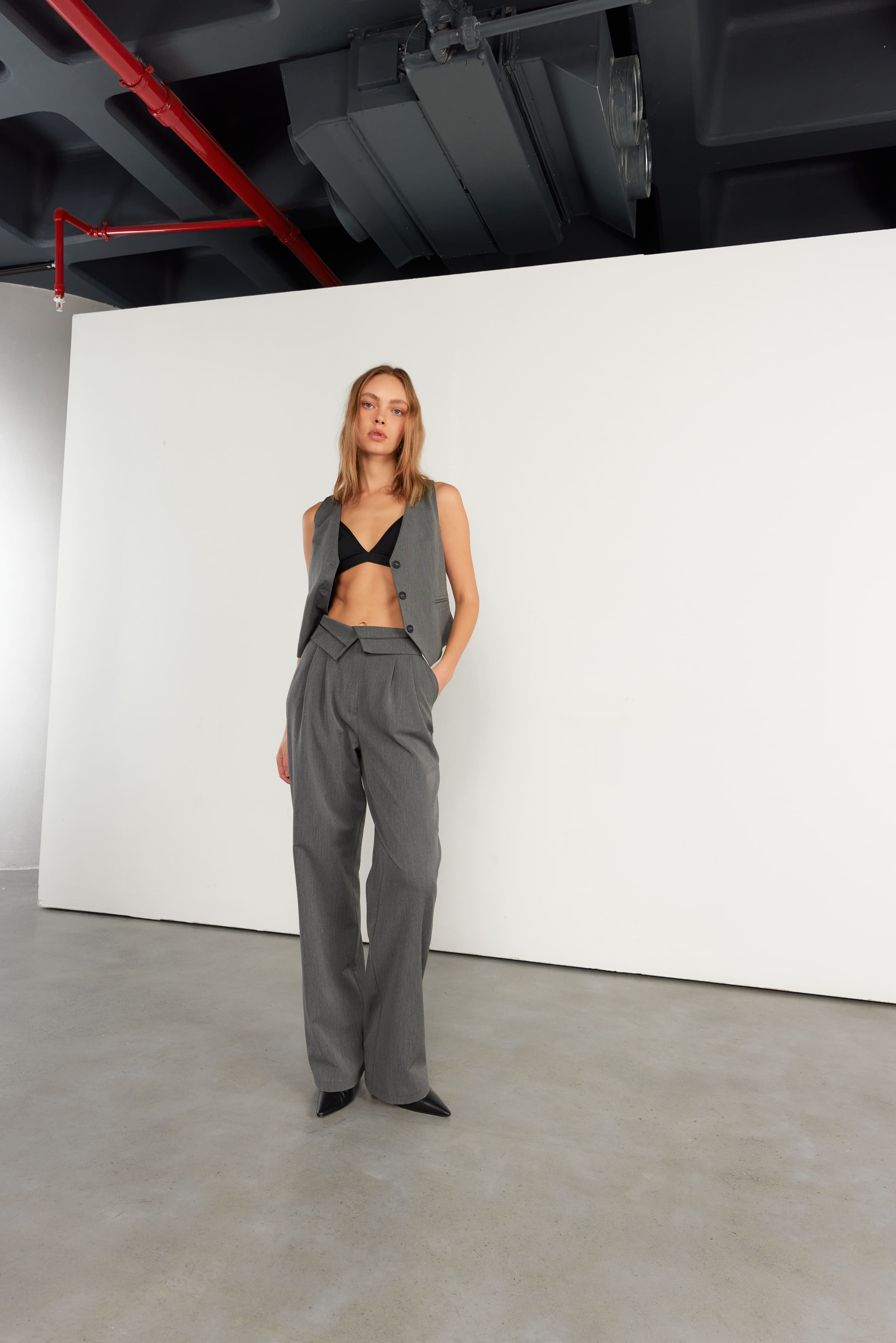A grey waistcoat left open over a black bralette with pleated trousers, set wide in Vatkalı's raw studio with its signal-red pipe.