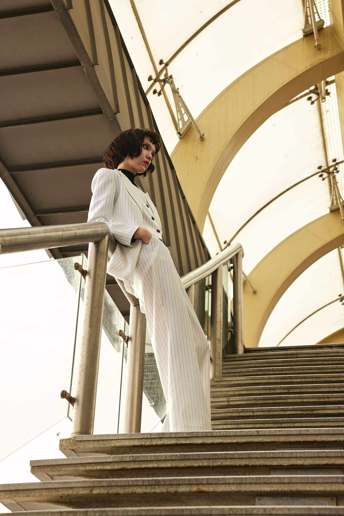 Shot from below, a white pinstripe Vatkalı suit leans against a staircase rail beneath vaulted canopy arches.