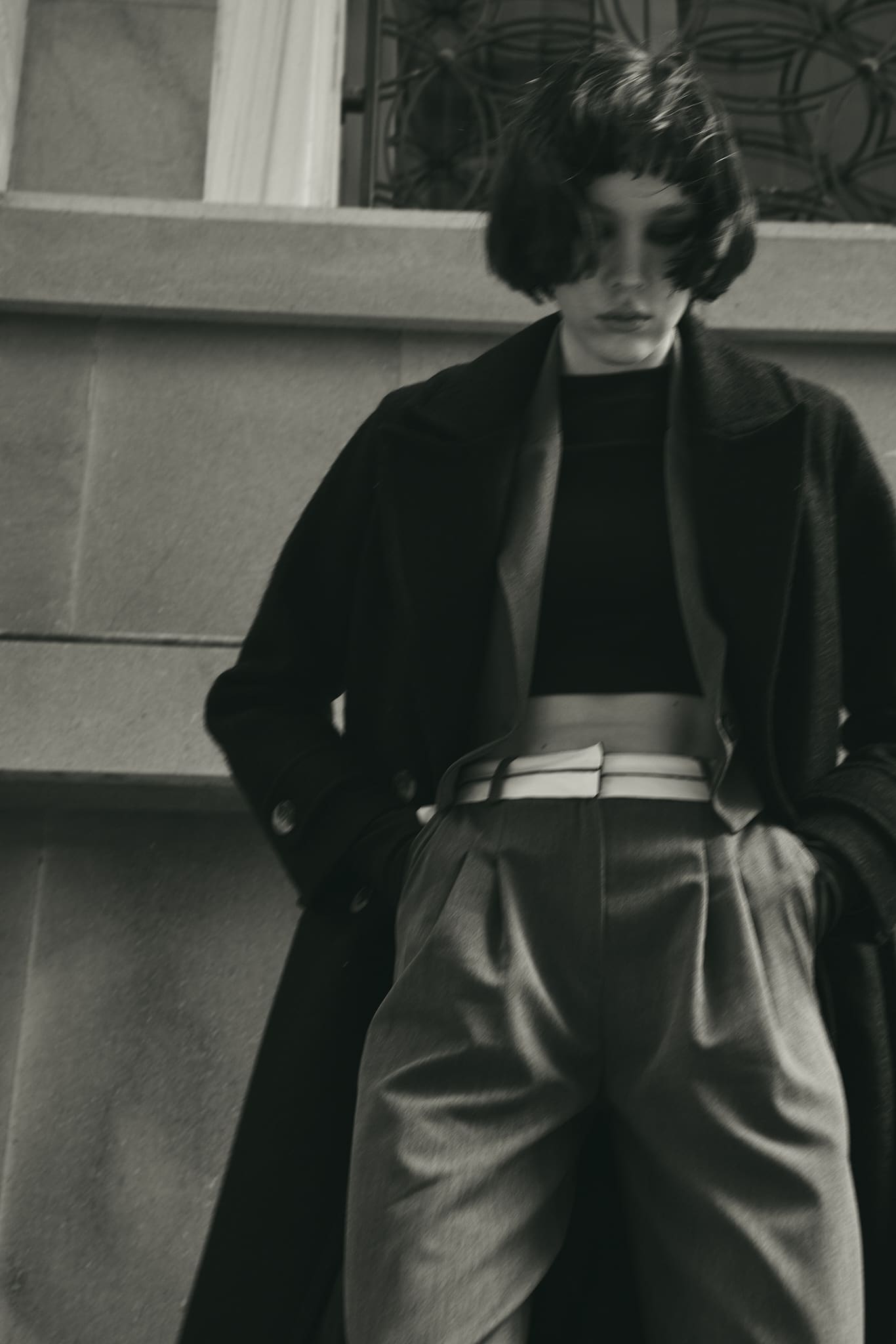 Hair sweeping across the face, an oversized dark coat opens over a contrast-belted high waist in a blurred monochrome Vatkalı frame.