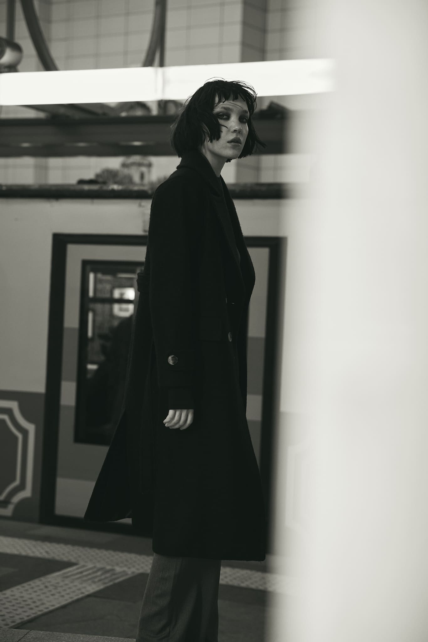 A long black Vatkalı coat glances back over the shoulder on a tram platform, framed past a soft foreground column in black and white.