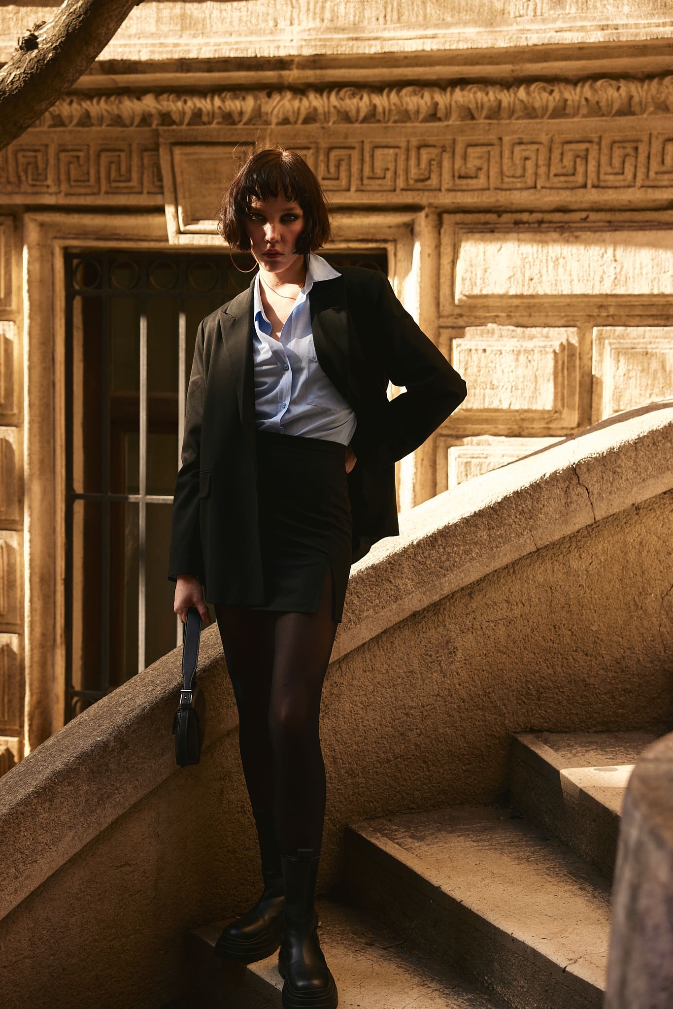 A black blazer and mini skirt over a sky-blue shirt descend a carved stone staircase in late sun for Vatkalı.