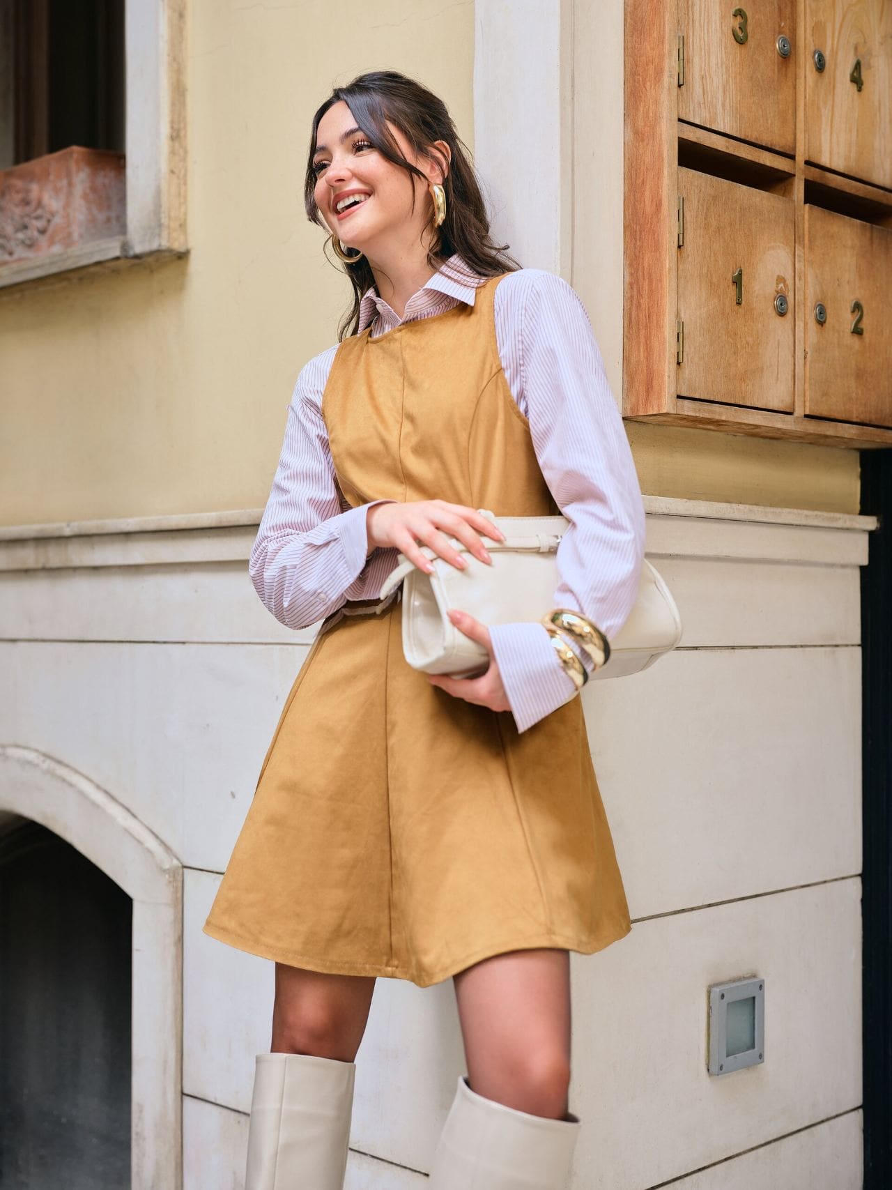 Derya Pınar Ak laughs beside vintage wooden mailboxes in a camel pinafore dress layered over a striped shirt, clutching a cream bag for XSIDE.