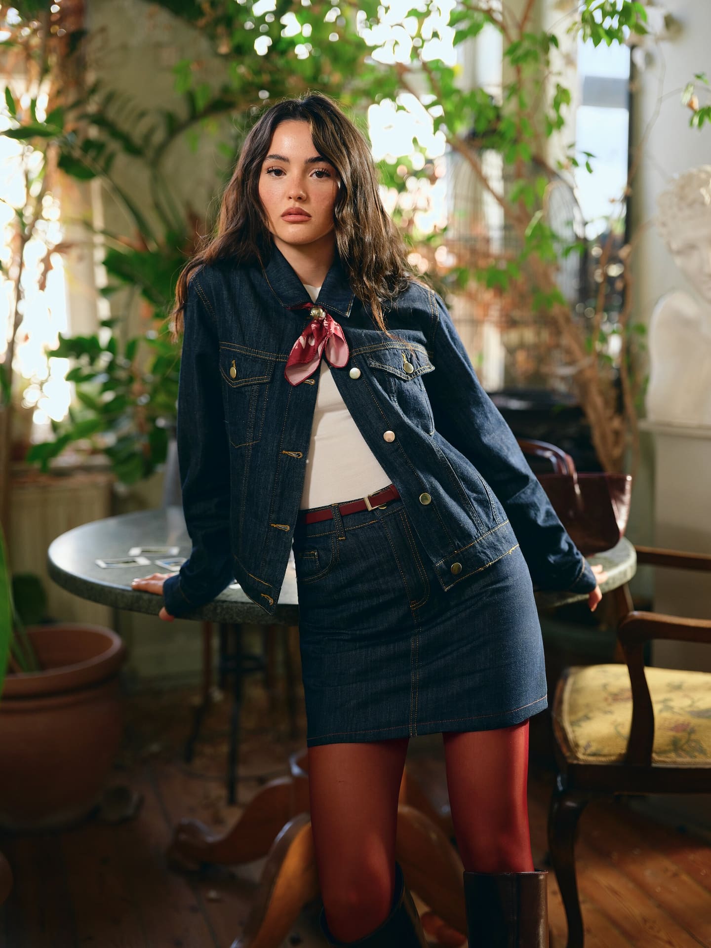 Derya Pınar Ak leans back on a marble bistro table in head-to-toe indigo denim with a red neck scarf, framed by winter-garden greenery for XSIDE.