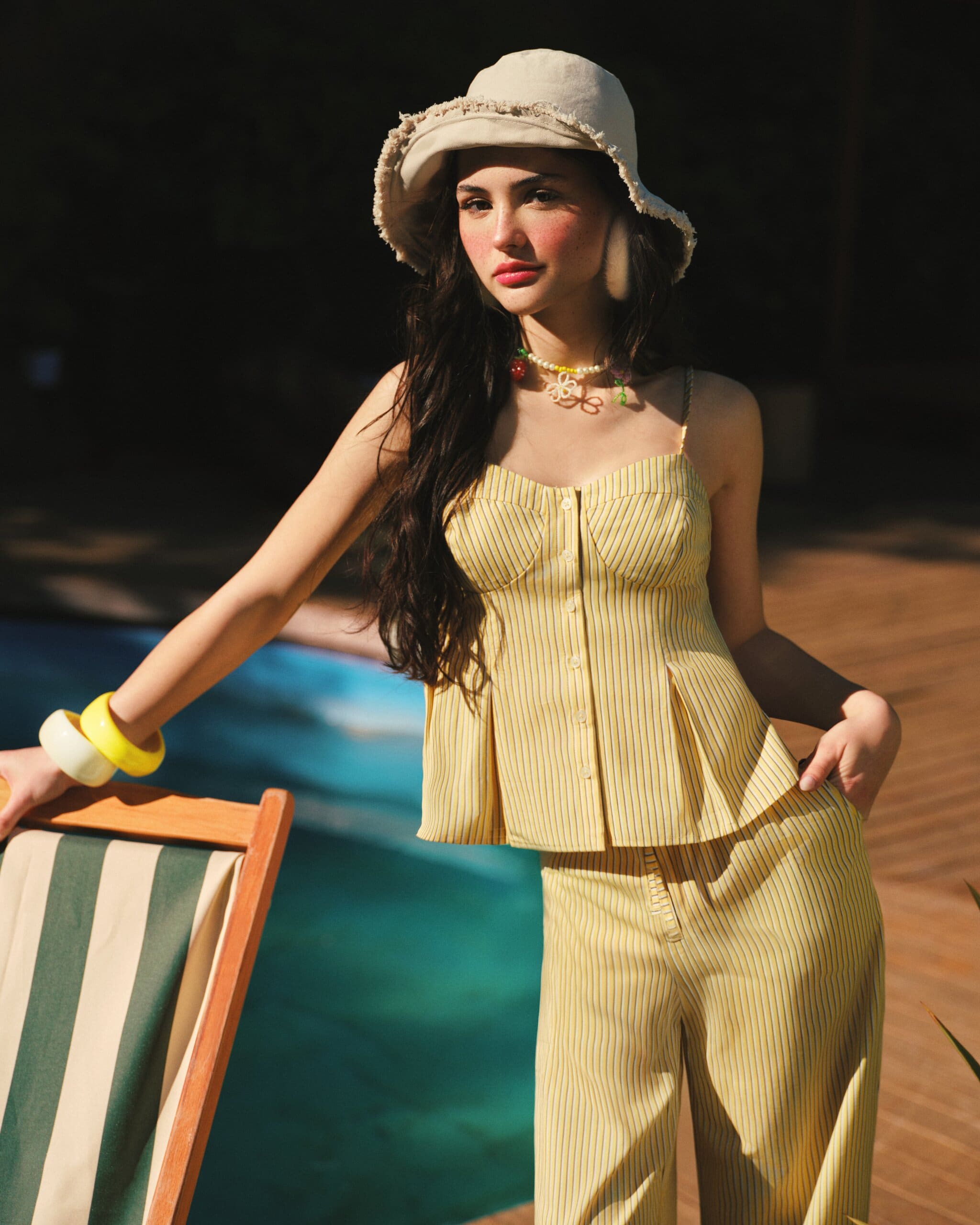 Derya Pınar Ak in a butter-yellow pinstripe bustier set and frayed bucket hat, resting a hand on a striped deck chair at the poolside for XSIDE.