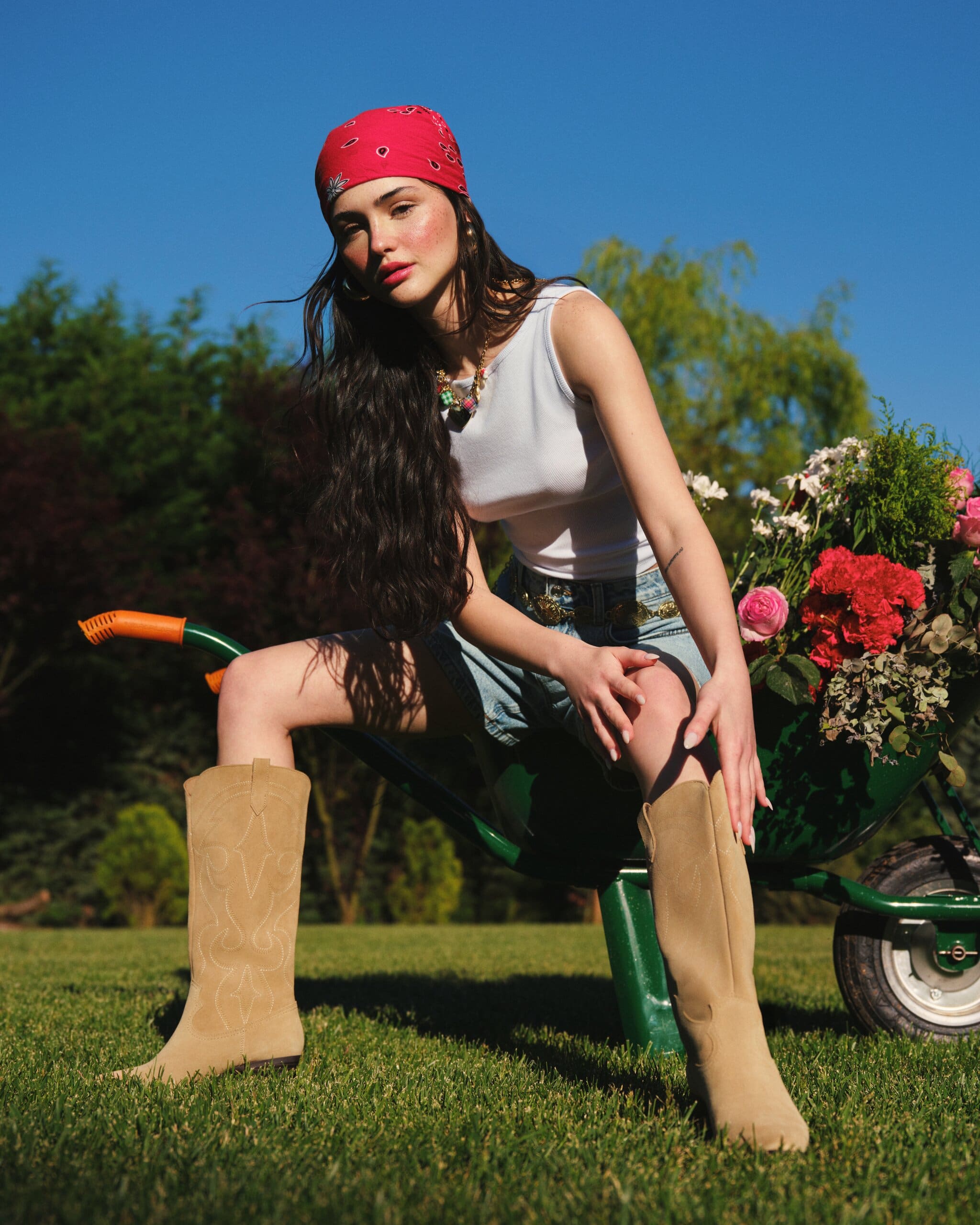Derya Pınar Ak perches on a flower-filled wheelbarrow in a red bandana, white tank and suede cowboy boots, shot low against a clear blue sky for XSIDE.